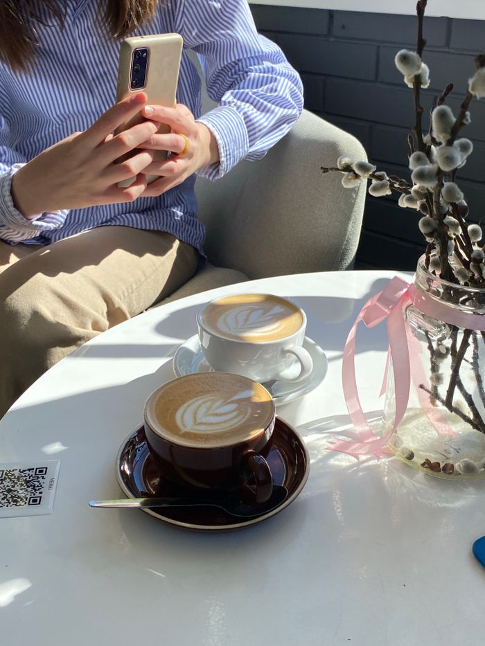 Woman photographing cappuccinos with latte art in a sunlit café scene.